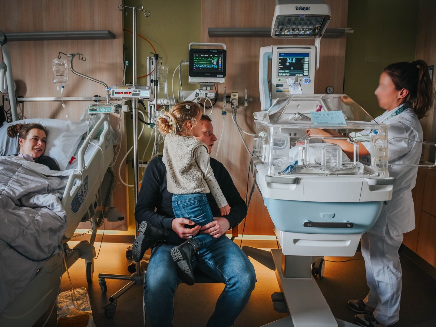 Parents beside premature newborn in hospital incubator, with nurse providing neonatal care in NICU ward.