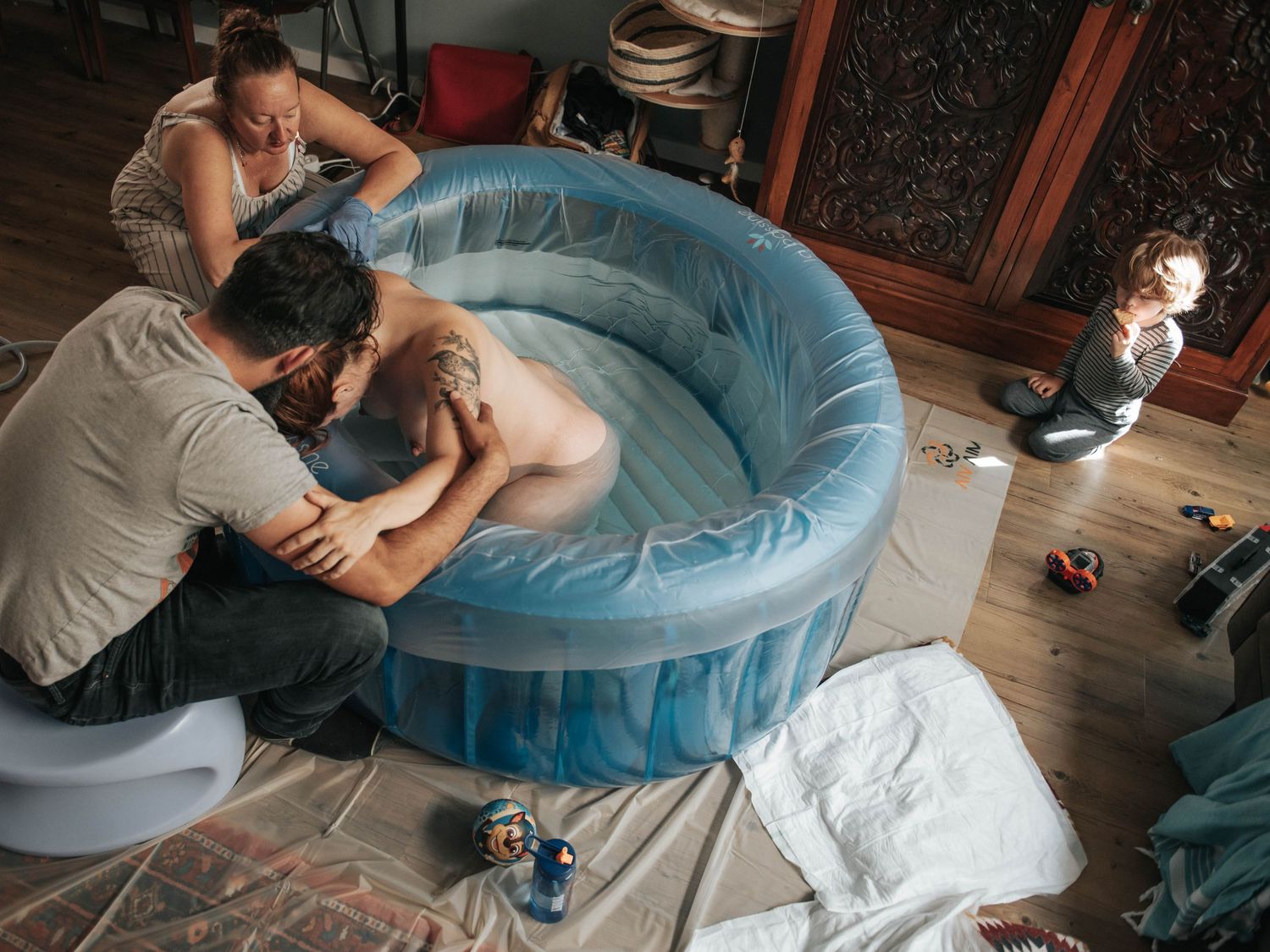 Family gathered around an inflatable birthing pool during a home water birth, with a toddler watching nearby.