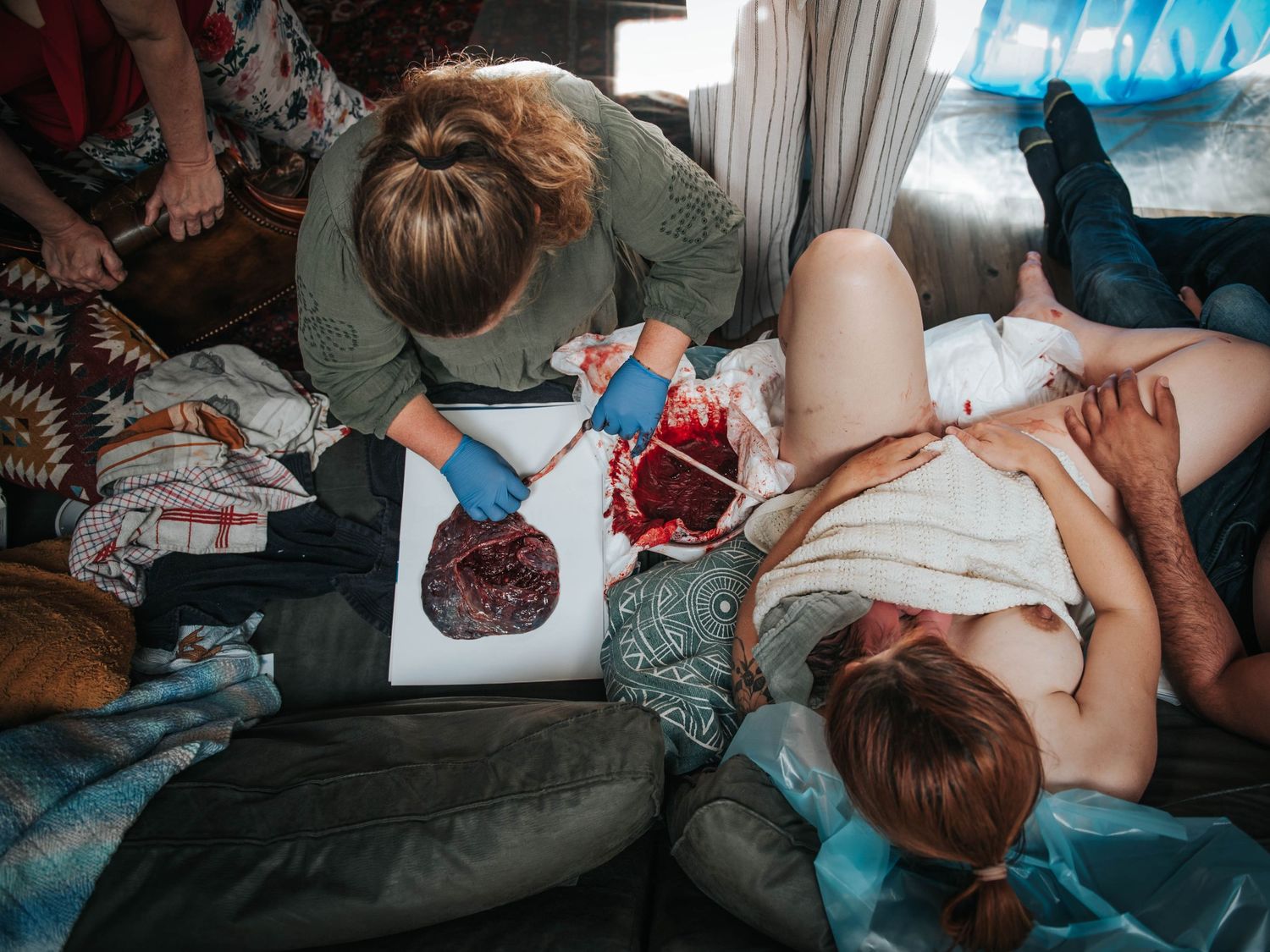 Midwife examining placenta after home birth while new mother rests on couch with support person nearby.