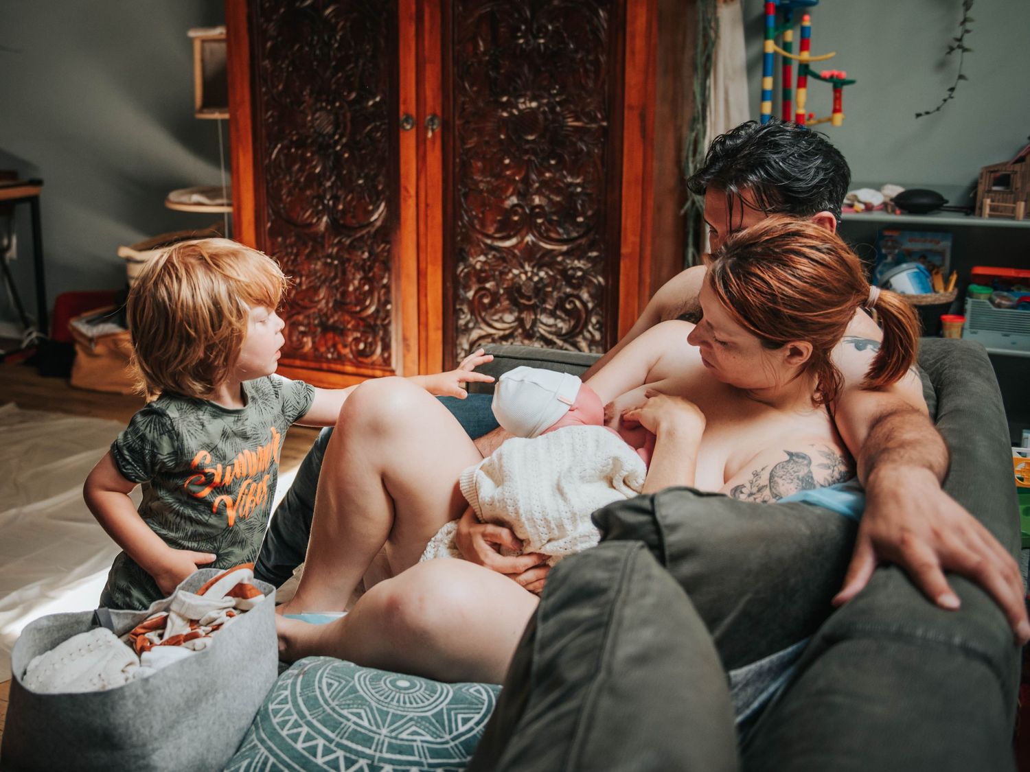 A mother breastfeeds a newborn on a couch while a toddler stands nearby in a cozy home setting.