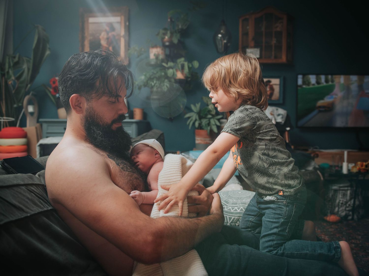 Bearded shirtless father holds newborn baby while young toddler child reaches over in a cozy plant-filled living room.