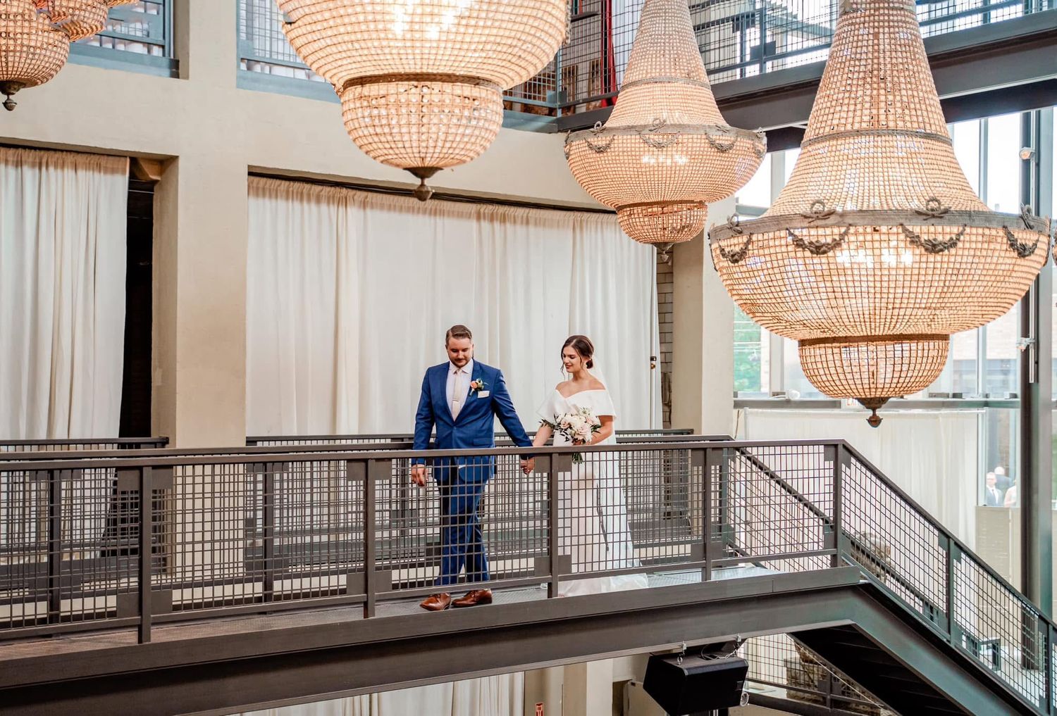 Bride and groom stand on elegant balcony surrounded by crystal chandeliers at a luxurious wedding venue.
