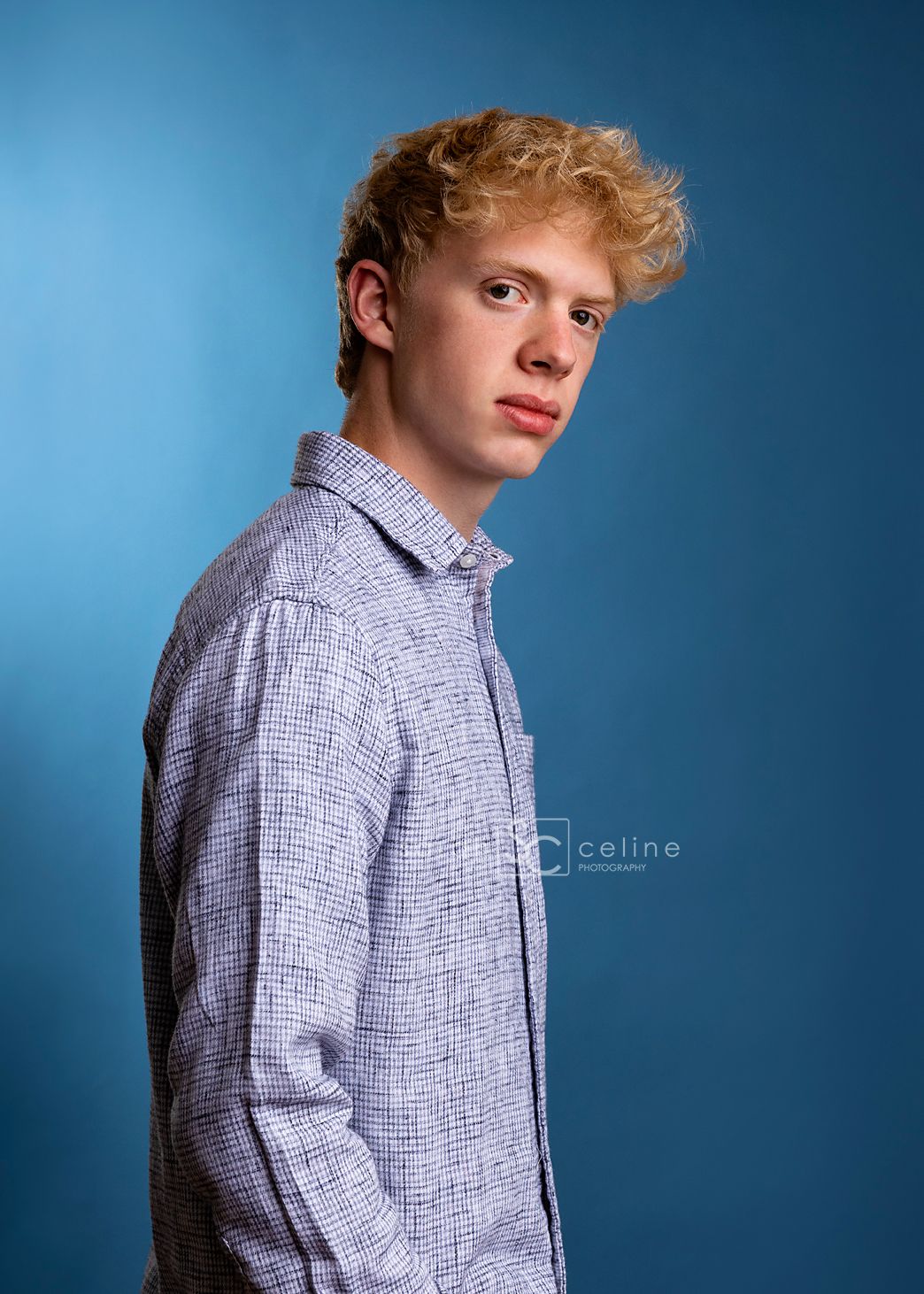 A young man with curly blonde hair poses in a blue patterned shirt against a blue studio background.