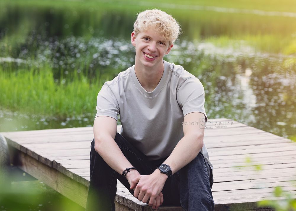 Smiling young blond man sitting on a wooden dock by a serene green pond on a sunny day.