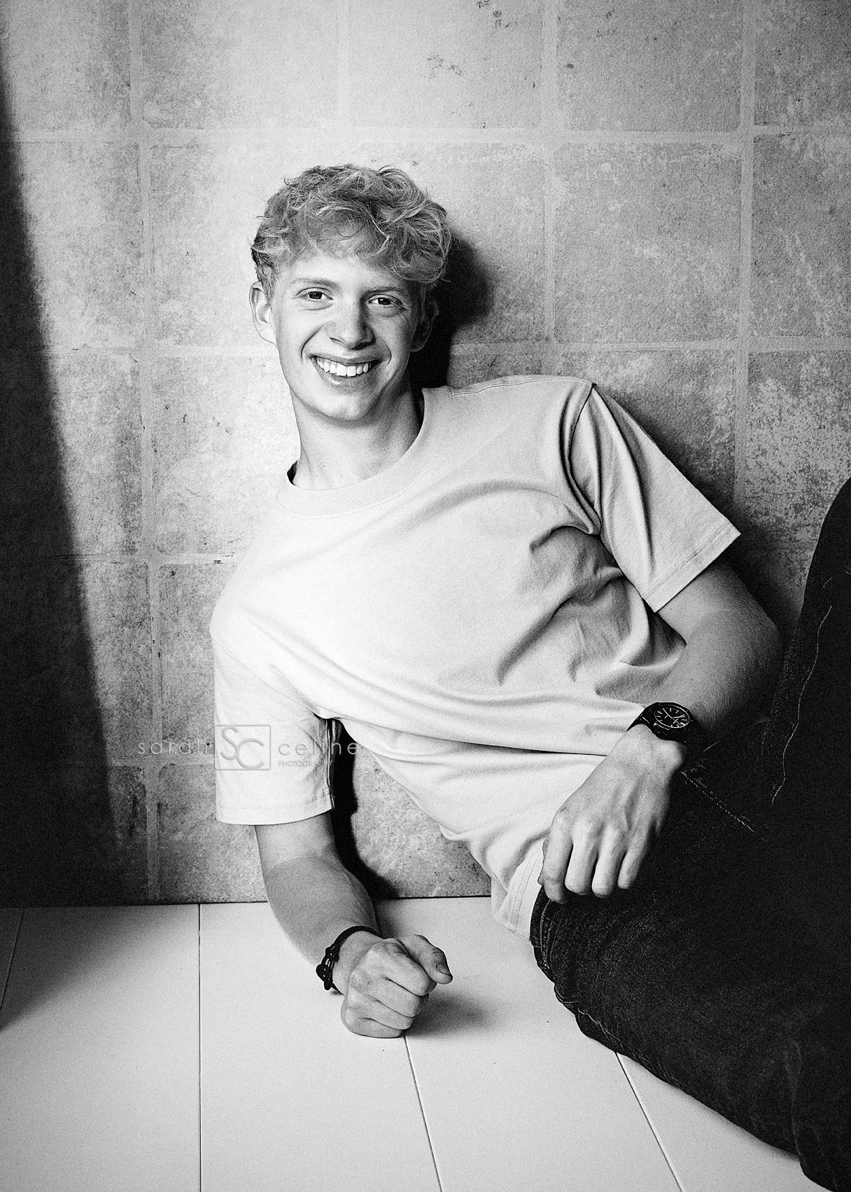 Smiling young man with curly hair sitting against a wall, wearing a t-shirt and jeans, black and white photo.