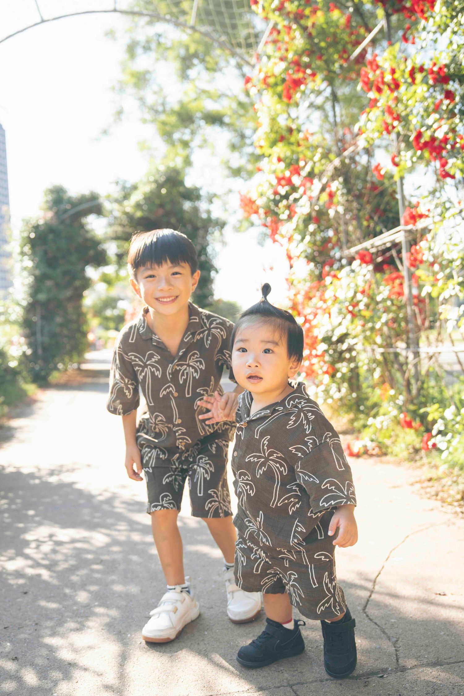 Two young Asian boys in matching brown palm tree print outfits smile outdoors near colorful red flowers.