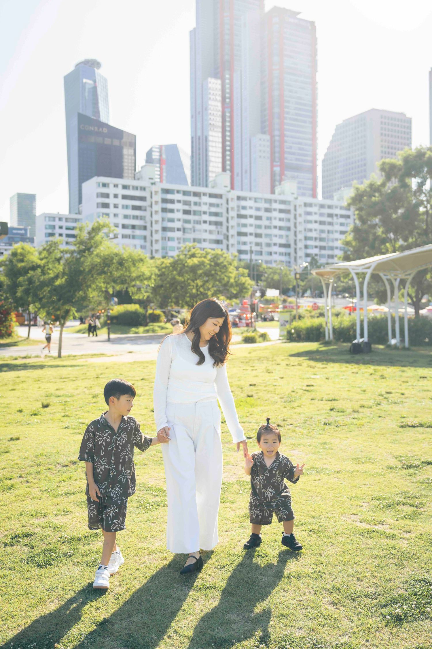A woman in white holds hands with two young boys in matching outfits in a sunny urban park with city skyline.