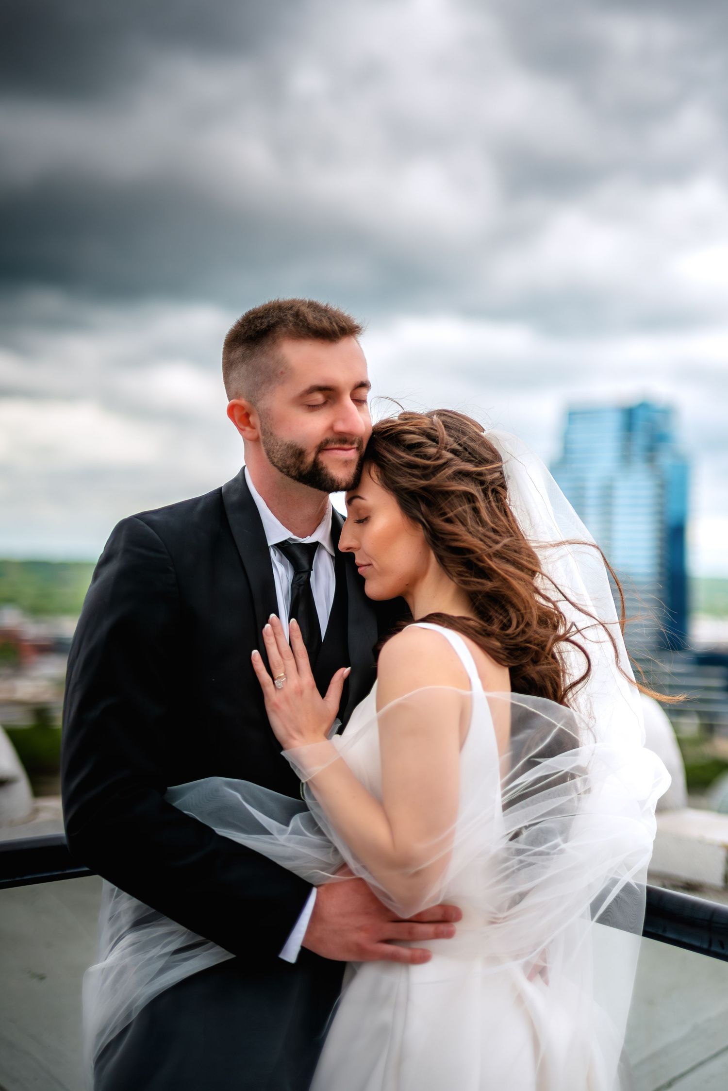 A bride and groom share a romantic embrace on a rooftop, with a dramatic cloudy sky and city skyline behind them.