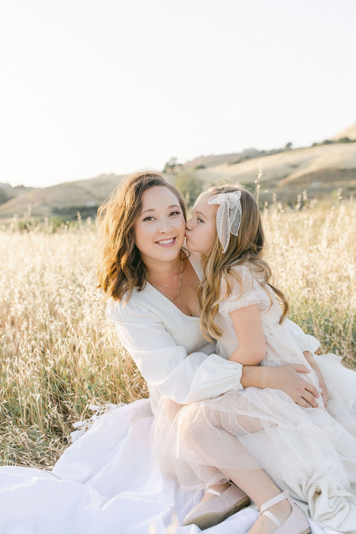 A smiling woman in white sits in a golden field as a young girl in a tulle dress gives her a sweet kiss on the cheek.