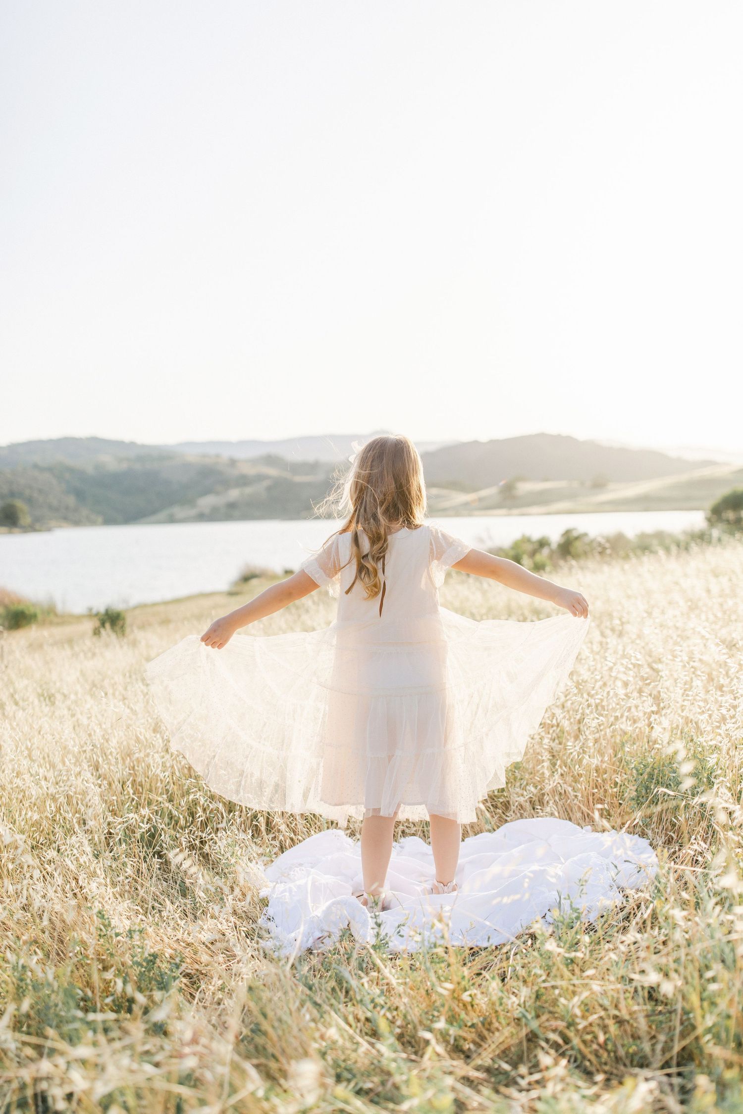 A young girl in a white dress stands in a golden field, arms outstretched, facing a serene lake and rolling hills.
