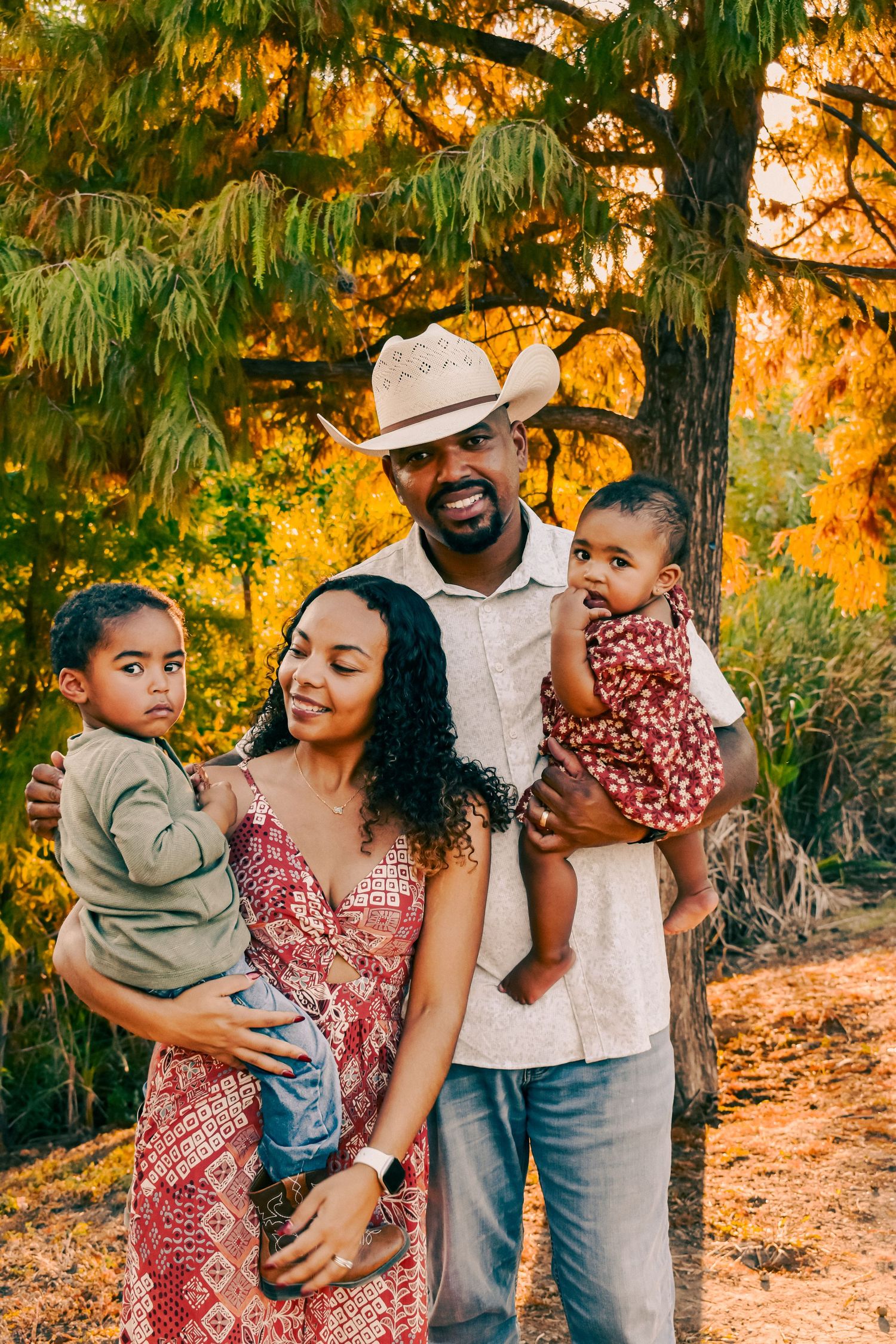 A family of four poses outdoors in golden hour light, parents holding two young children near autumn-colored trees.