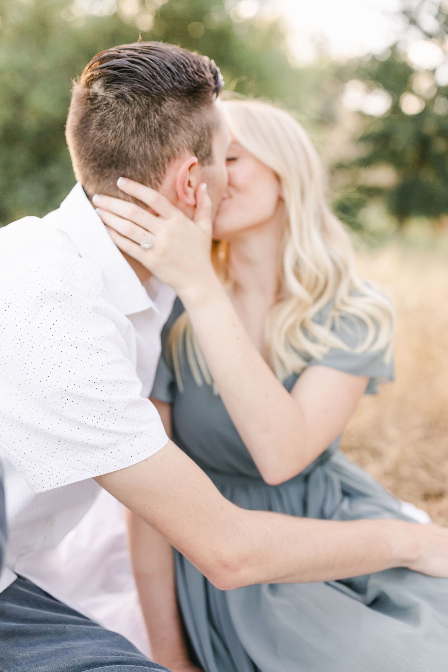 Couple sharing a romantic kiss outdoors, woman in sage dress touching man's face, soft natural light background.