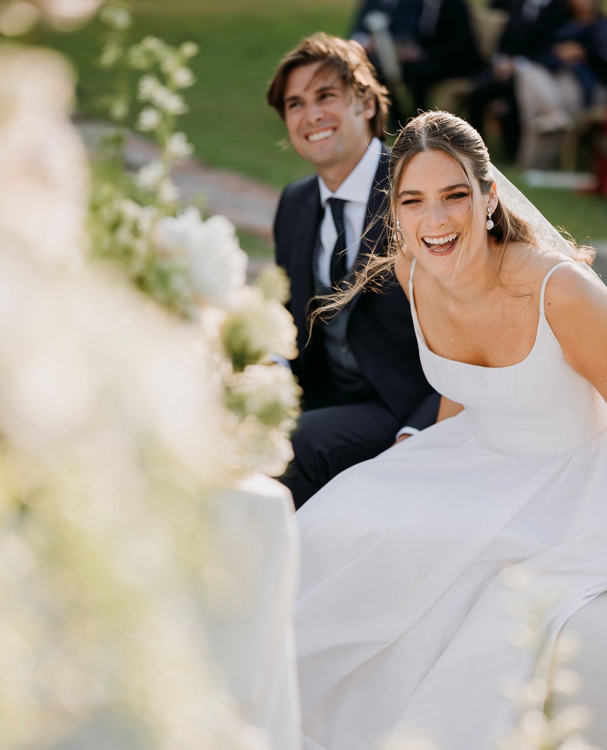 Laughing bride in white gown seated with groom during outdoor wedding ceremony with floral decor.
