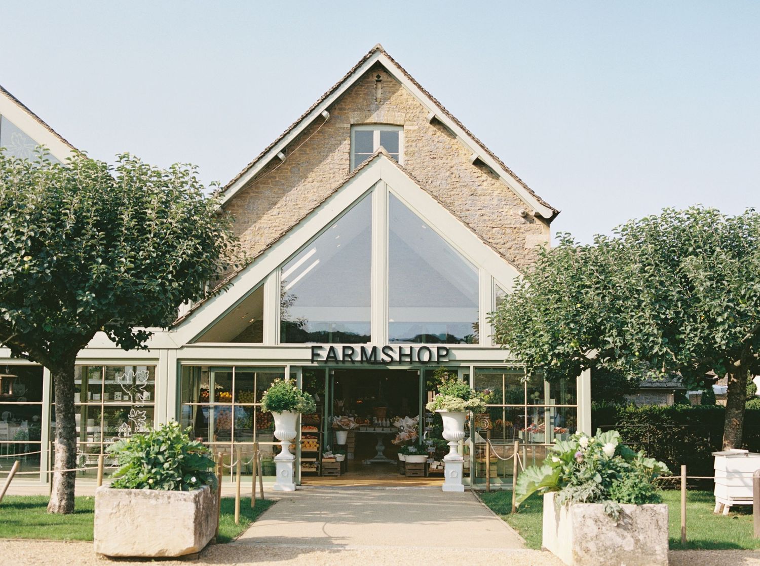 Rustic farmshop building with large glass facade, brick walls, and lush greenery on a sunny day.