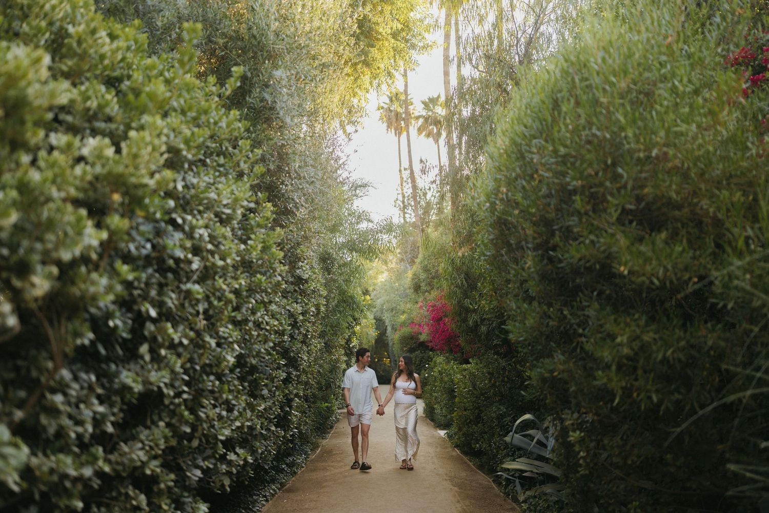 Couple holding hands walking down a lush garden path lined with tall green hedges and trees.