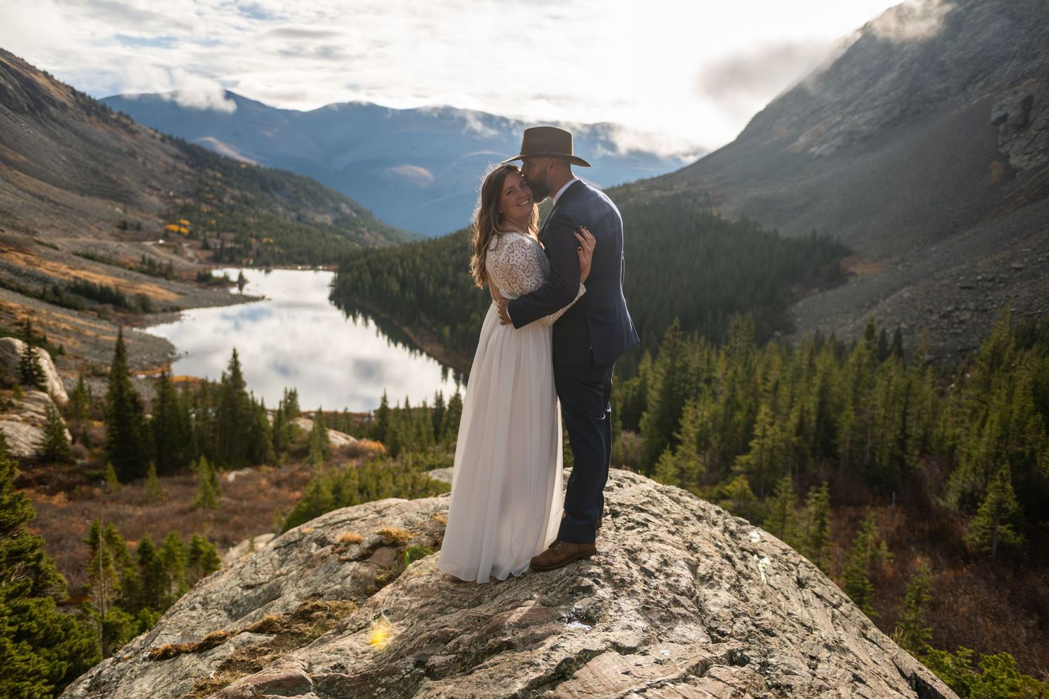 Couple shares a romantic kiss on a rocky summit with a stunning mountain lake and forest valley backdrop.