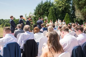 Outdoor wedding ceremony with guests seated in white chairs facing groom and officiant beneath floral arch on sunny day.
