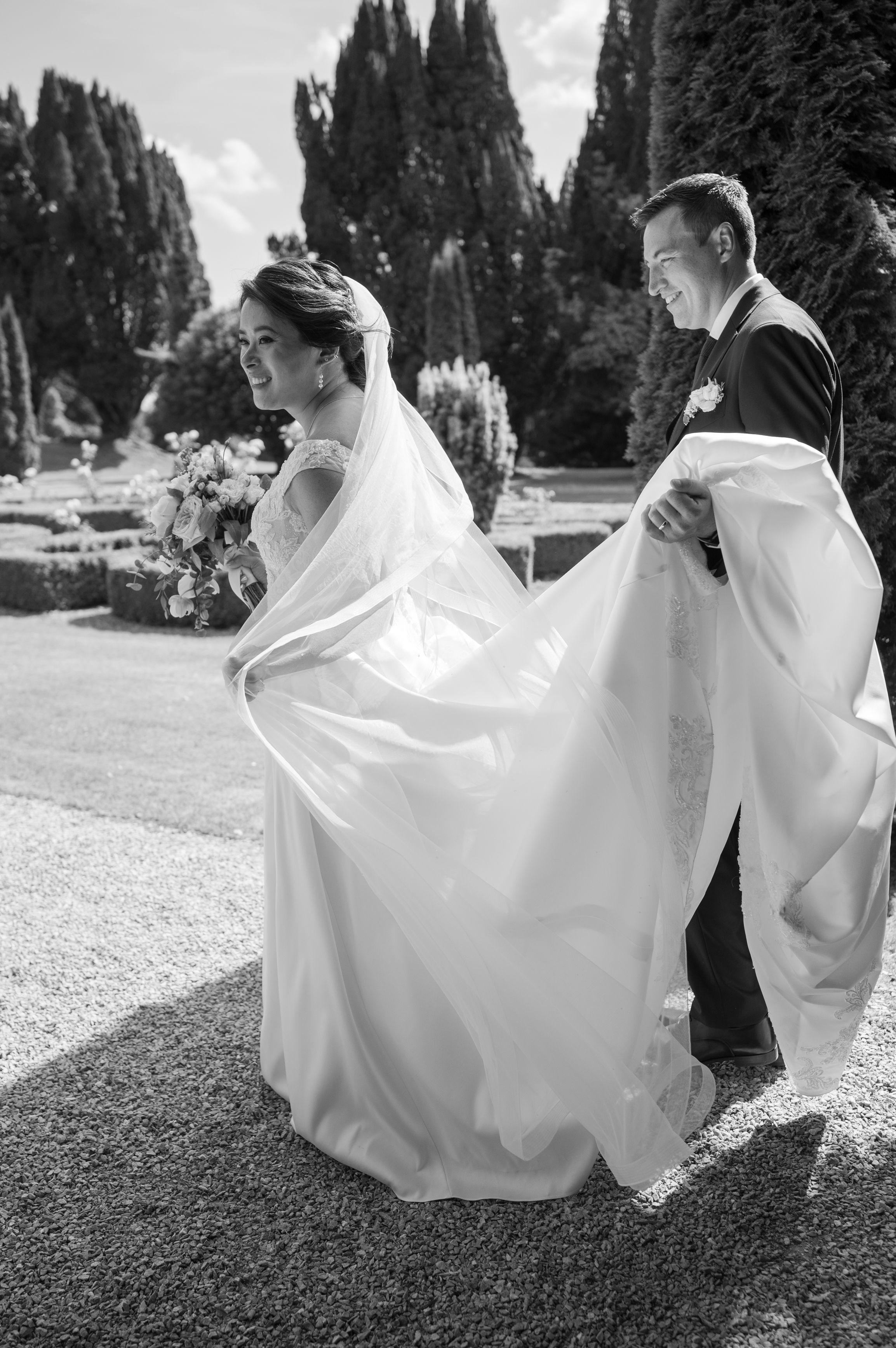 Black and white photo of a bride holding bouquet while groom lifts her wedding dress train in a garden.