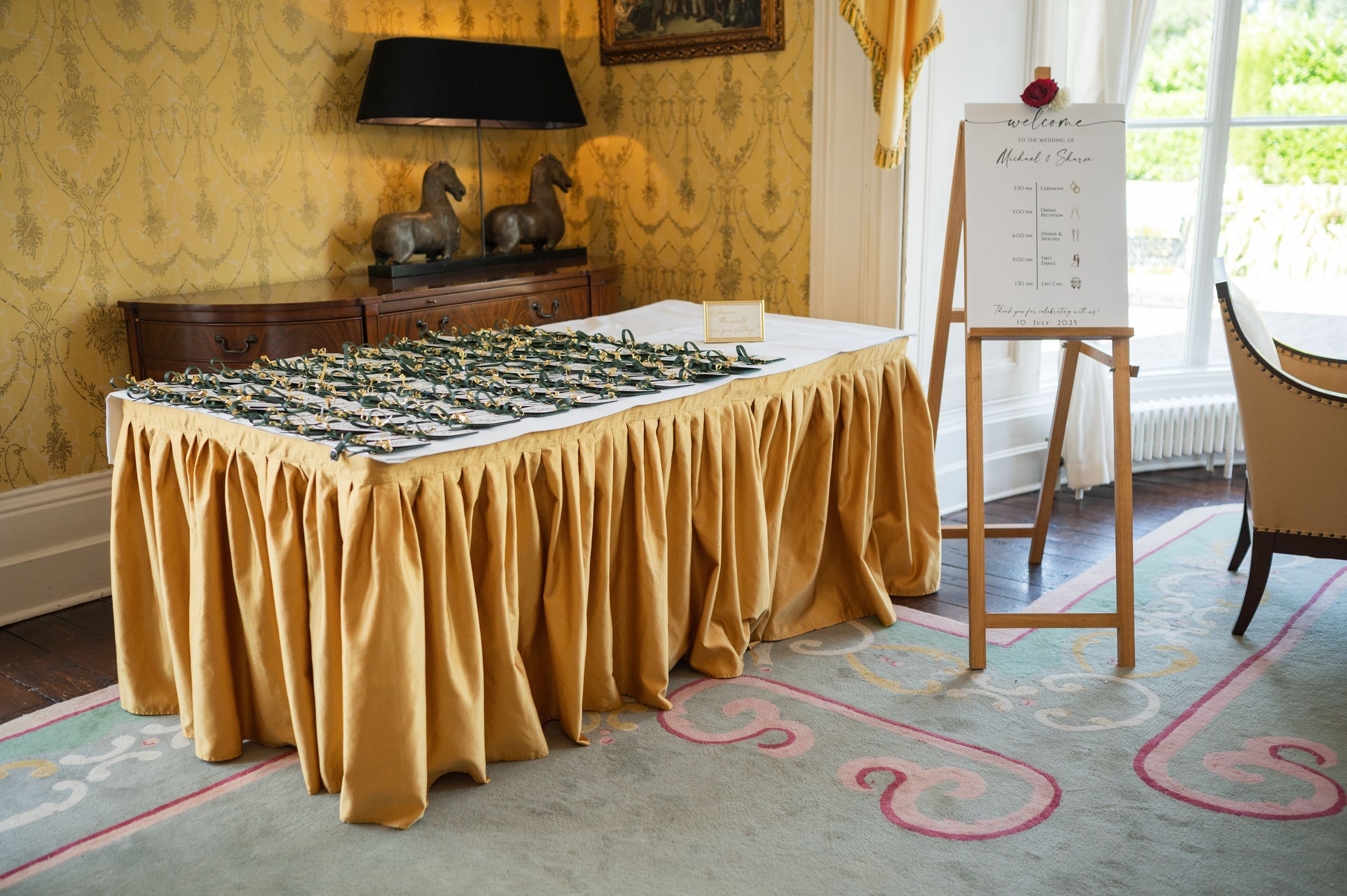 Elegant gold skirted table displaying wedding seating chart cards beside a wooden easel in ornate room.