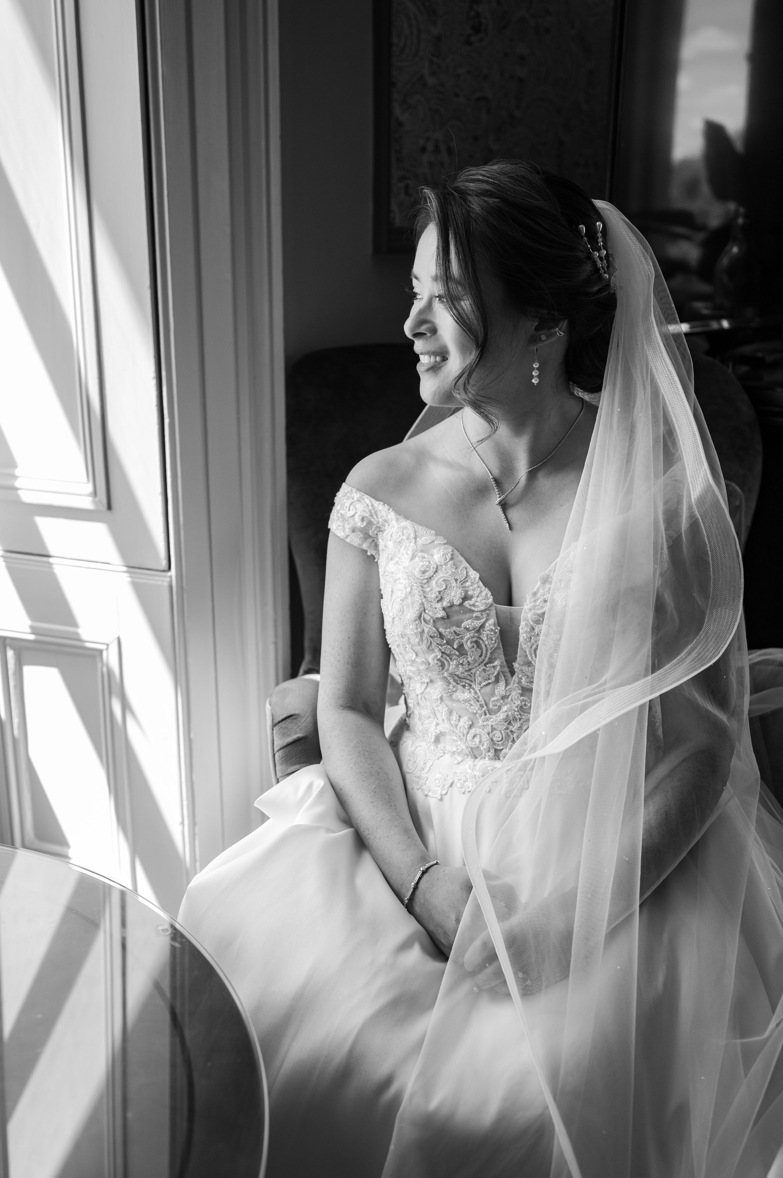 Black and white photo of a smiling bride in lace gown and veil sitting by a sunlit window on her wedding day.