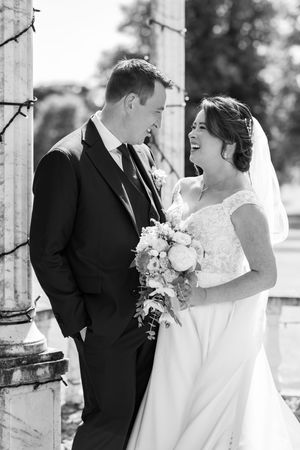Black and white photo of a laughing bride holding a bouquet and groom in a suit standing by stone columns.