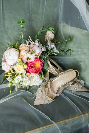 Bridal bouquet with pink peonies and roses beside elegant lace wedding heels on a green velvet veil.