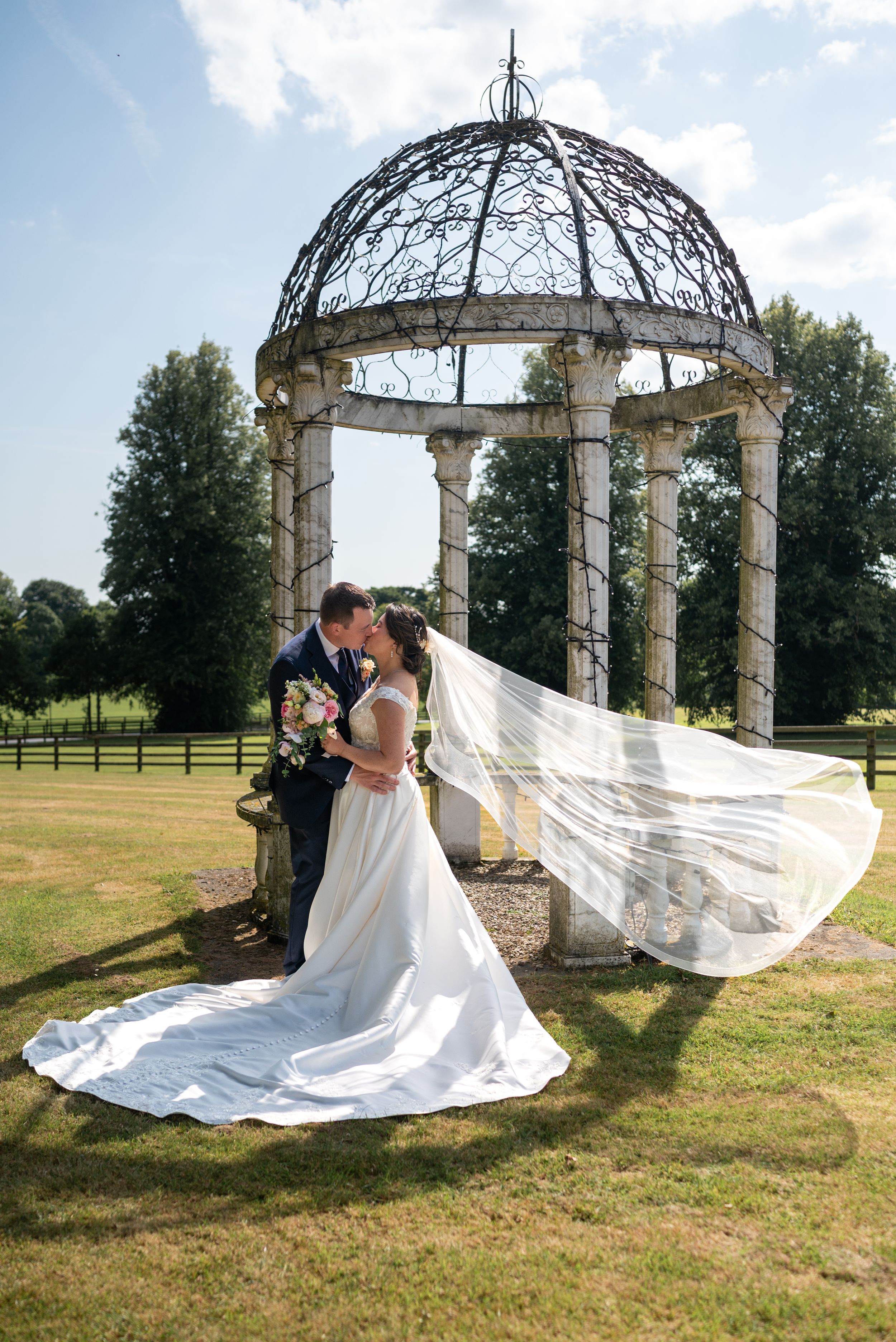 Bride and groom kissing under ornate stone gazebo, wedding veil flowing in breeze, lush green garden setting on sunny day.