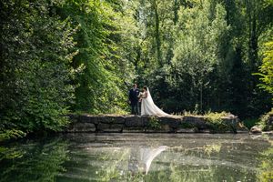 Bride and groom standing on stone bridge surrounded by lush green trees reflected in calm pond below.