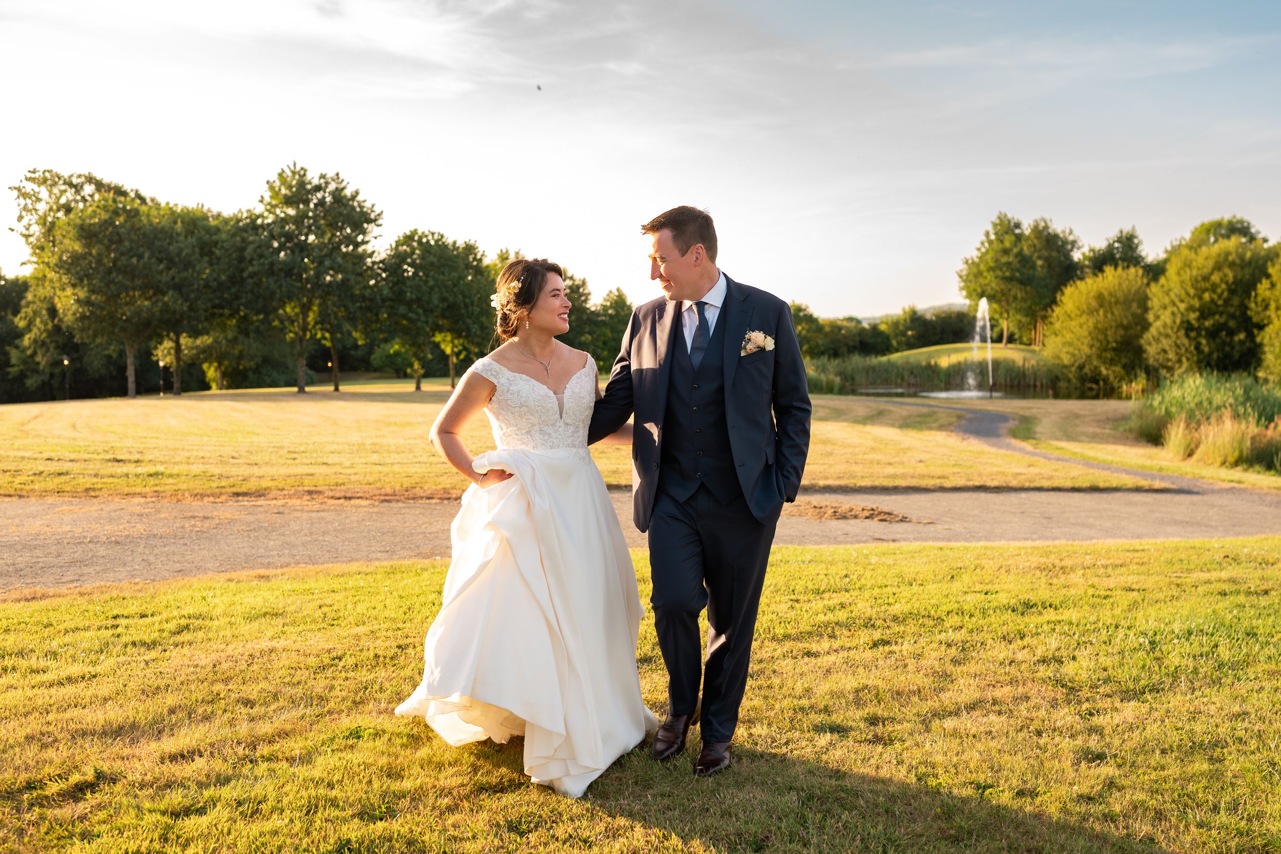 Bride and groom walk together at golden hour across lush grounds with a pond in the background.