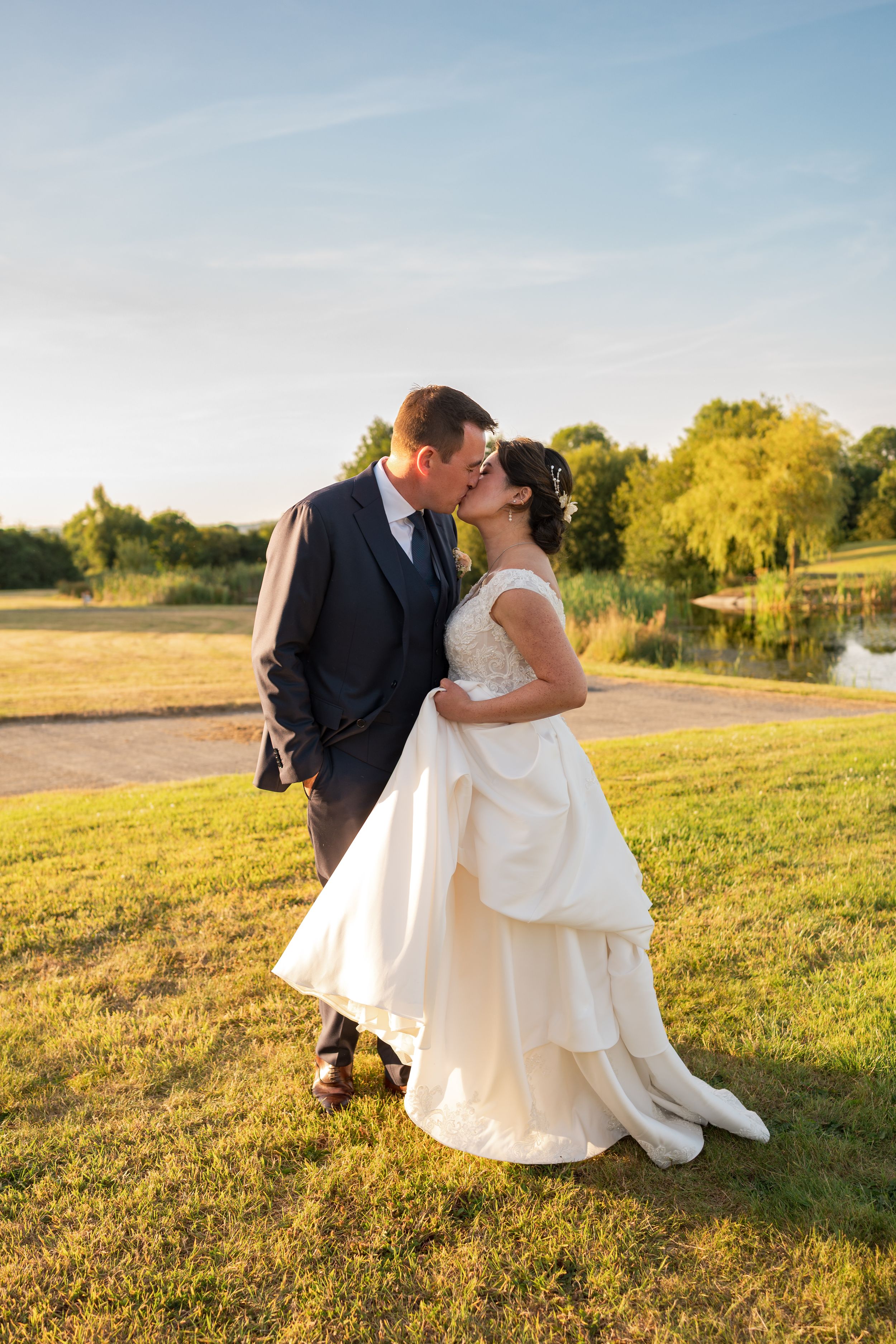 A bride and groom share a romantic kiss on a sunlit lawn with a serene lake and lush trees in the background.