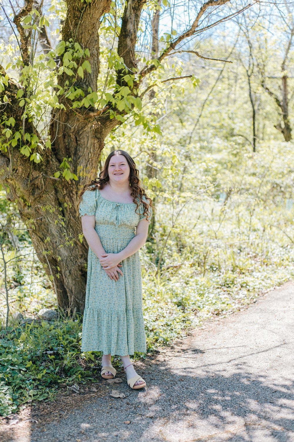 Young woman in green floral midi dress smiling beside a tree on a sunny spring day outdoors.
