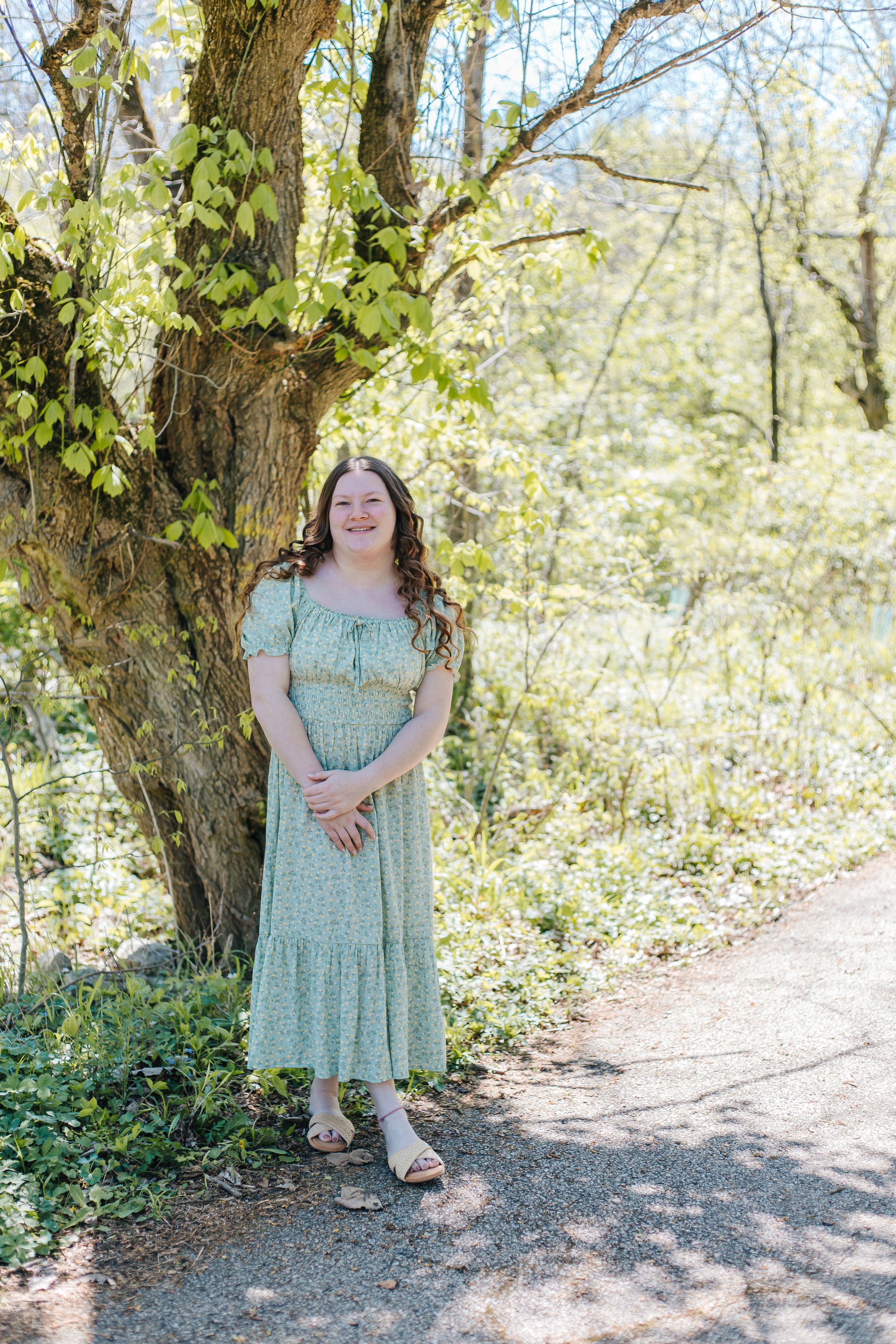Young woman in green floral midi dress smiling beside a tree on a sunny spring day outdoors.