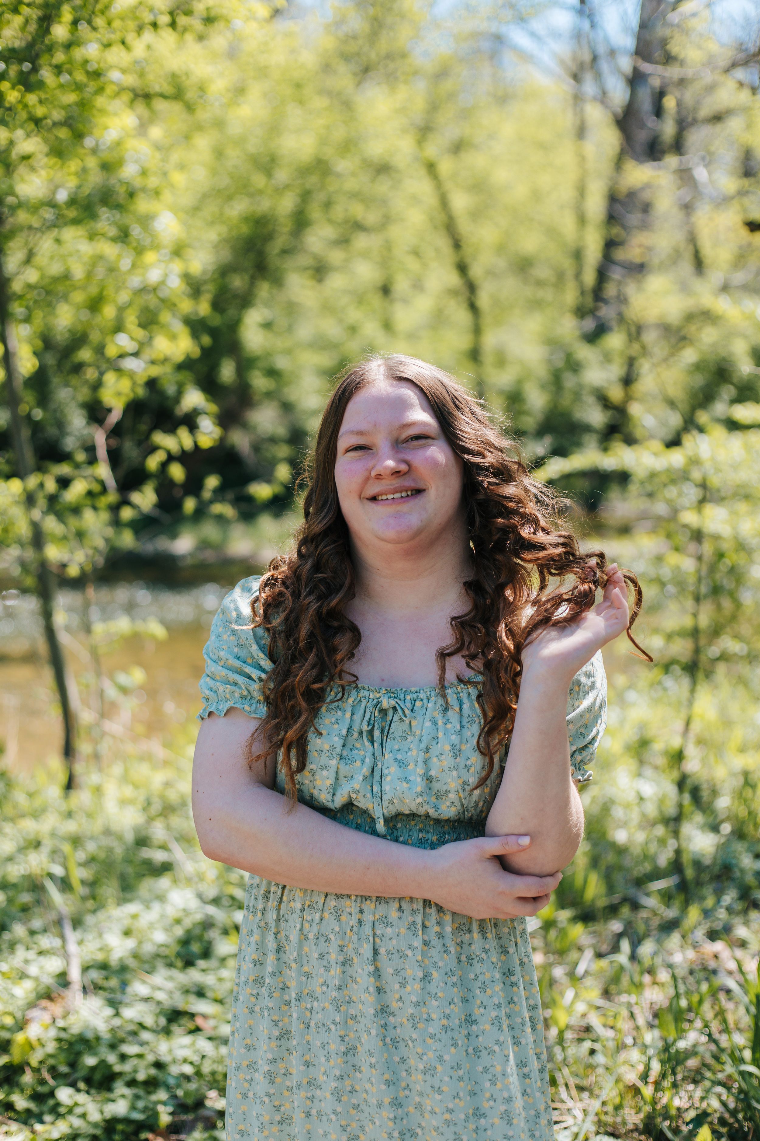 Young woman in floral off-shoulder dress smiling outdoors among lush green trees near a pond.