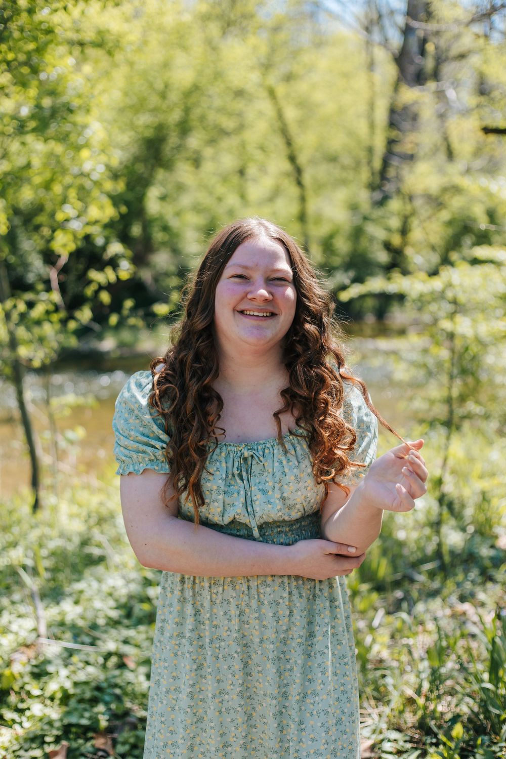 Smiling young woman with curly hair in a floral green dress standing outdoors in a lush green forest setting.
