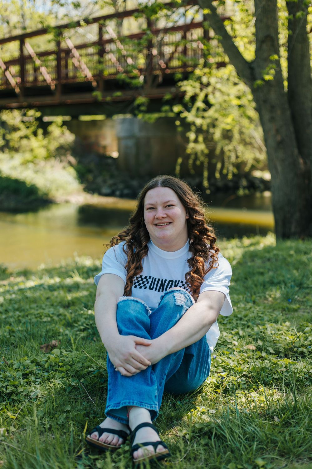 Young woman with curly hair sits smiling on green grass near a creek and bridge on a sunny day.