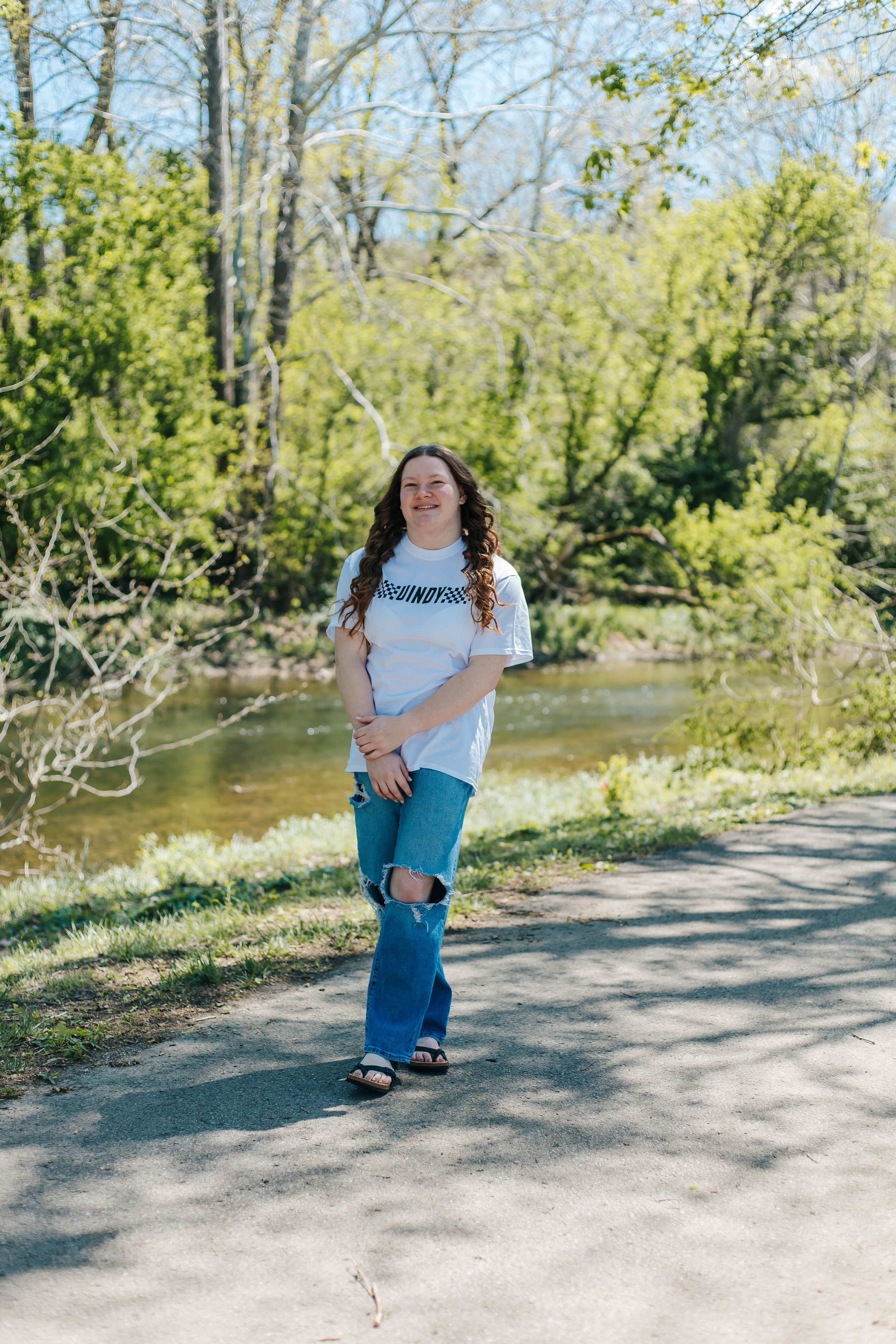 Smiling young woman in white tee and ripped jeans standing on a sunny park path near a river.