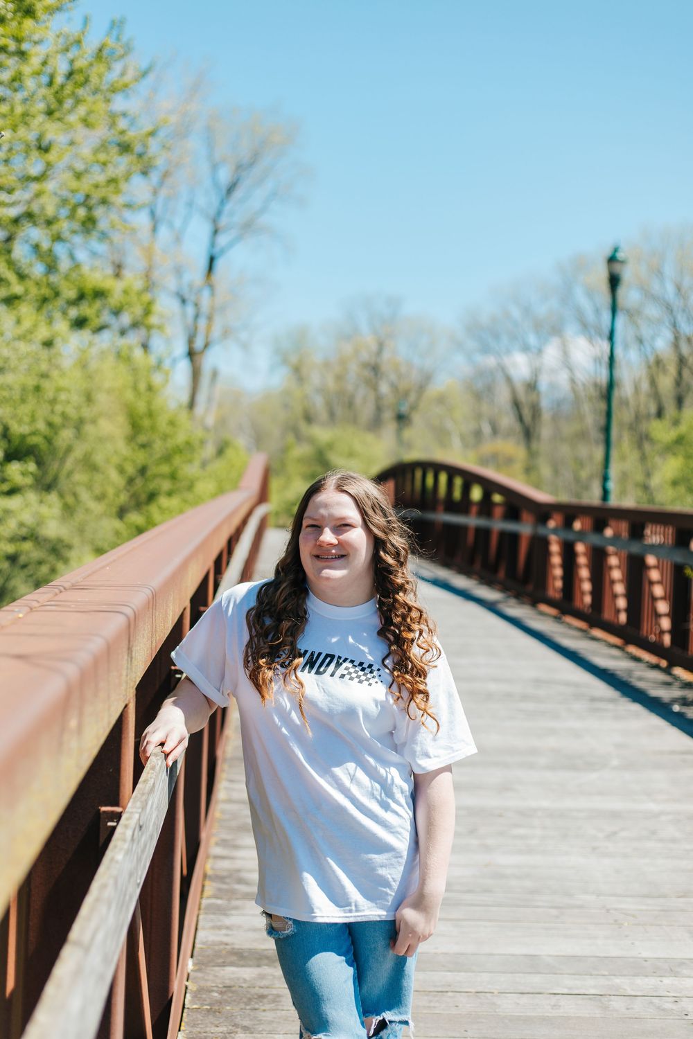 Young woman in white graphic tee leaning on a rustic bridge railing on a bright sunny day.