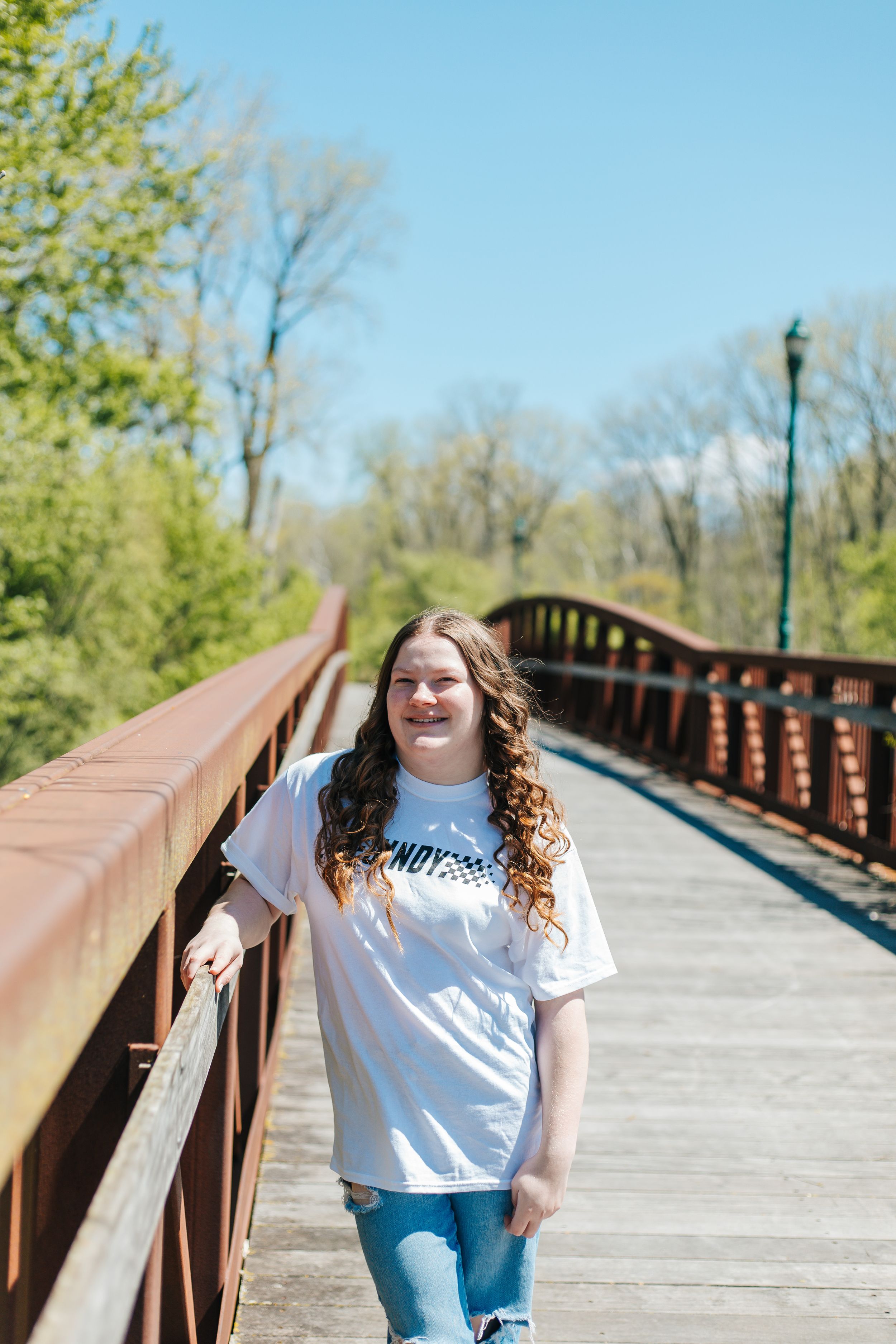 Young woman in white graphic tee leaning on a rustic bridge railing on a bright sunny day.