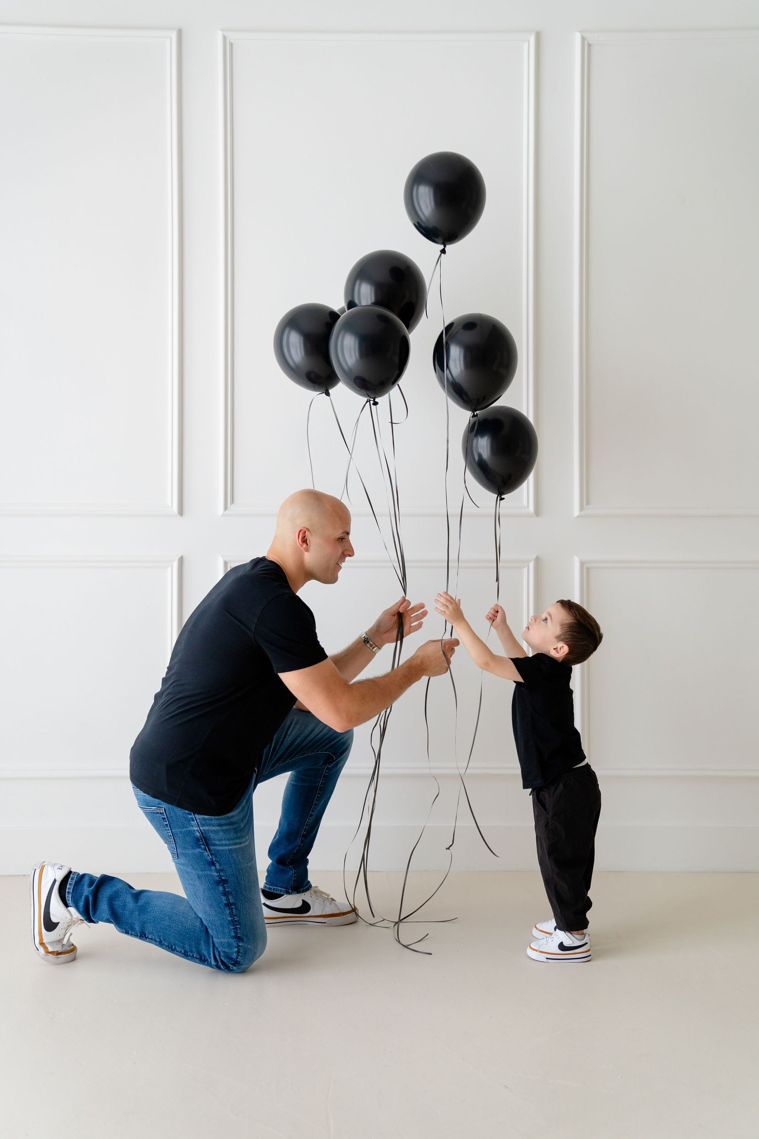 A bald man kneeling to hand black balloons to a young boy in a white studio setting.