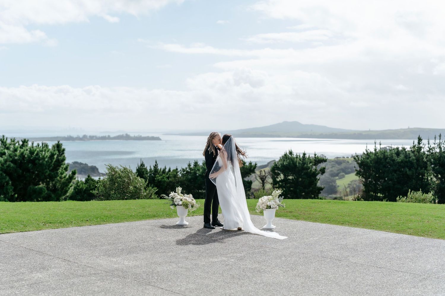 Bride and groom share a kiss at an outdoor wedding ceremony with stunning ocean and island views in the background.