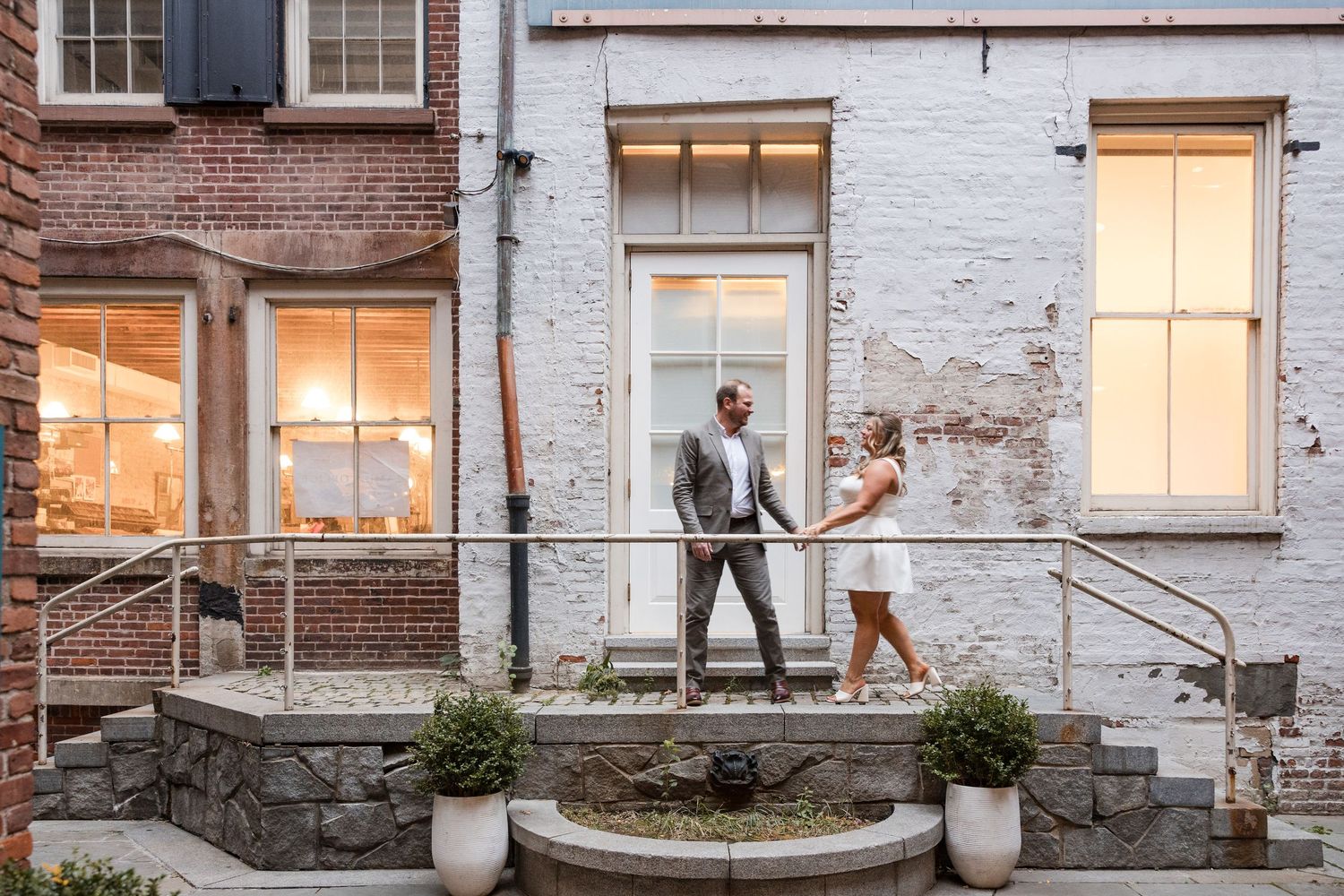 Joyful newlyweds hold hands as groom jumps mid-air on cobblestone street with city skyline backdrop.