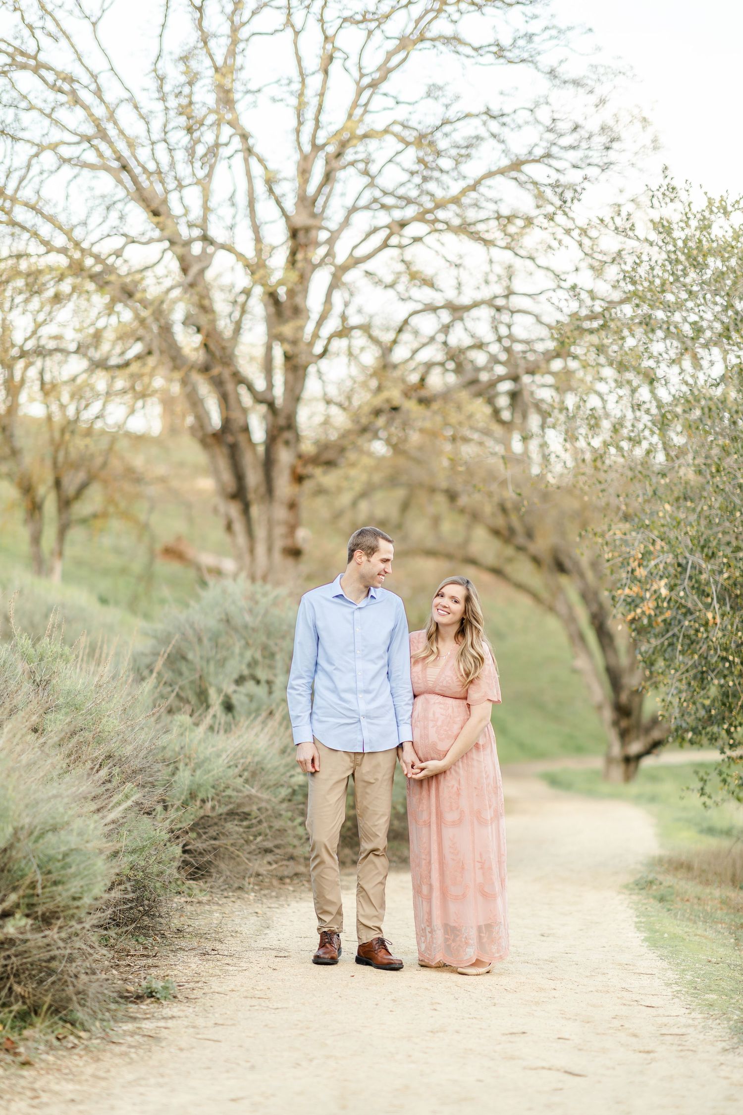 Couple holding hands on a scenic dirt path, woman in pink maternity dress, man in blue shirt, surrounded by trees.