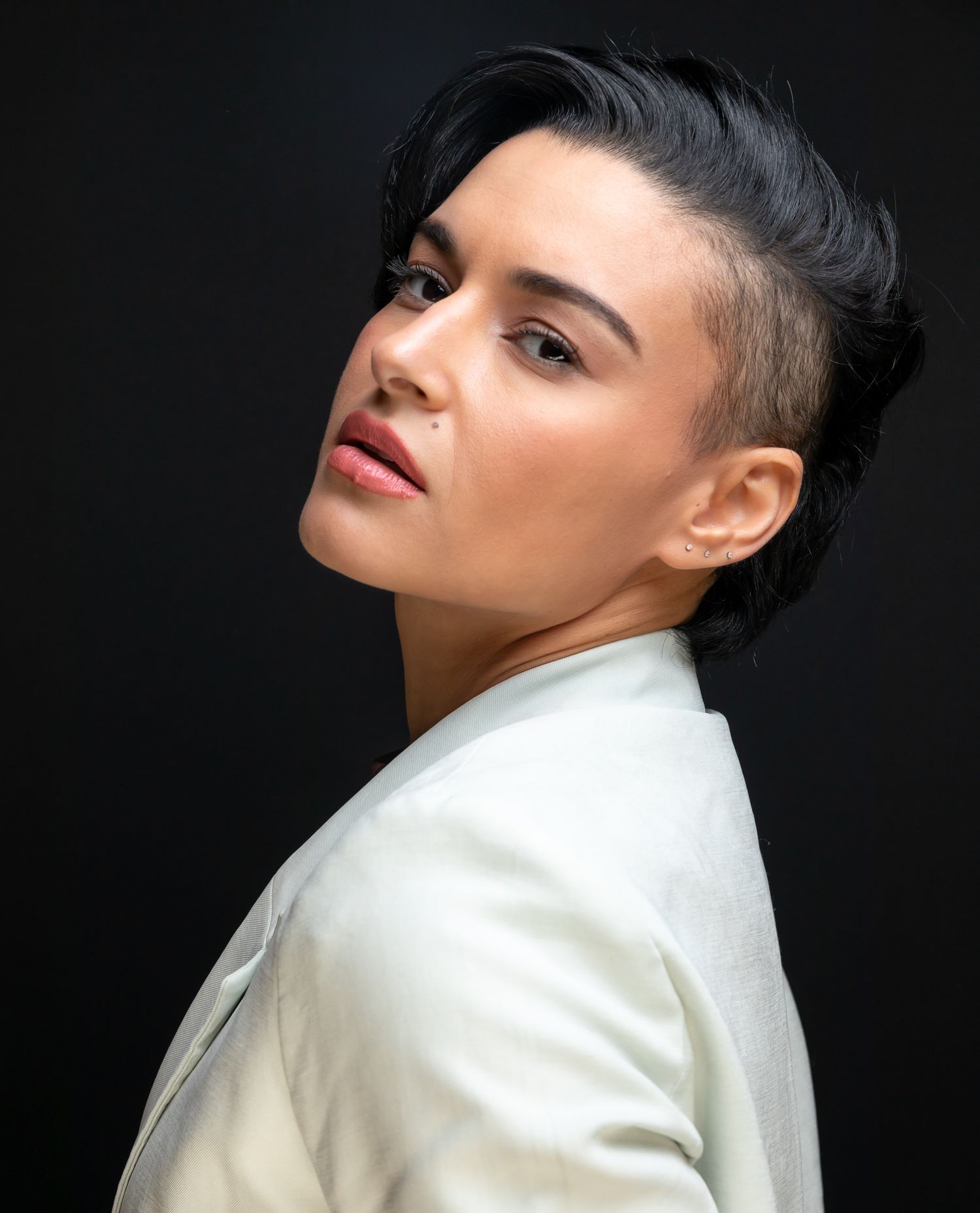 Portrait of a woman with shaved sides and dark upswept hair wearing a white blazer against a dark background.