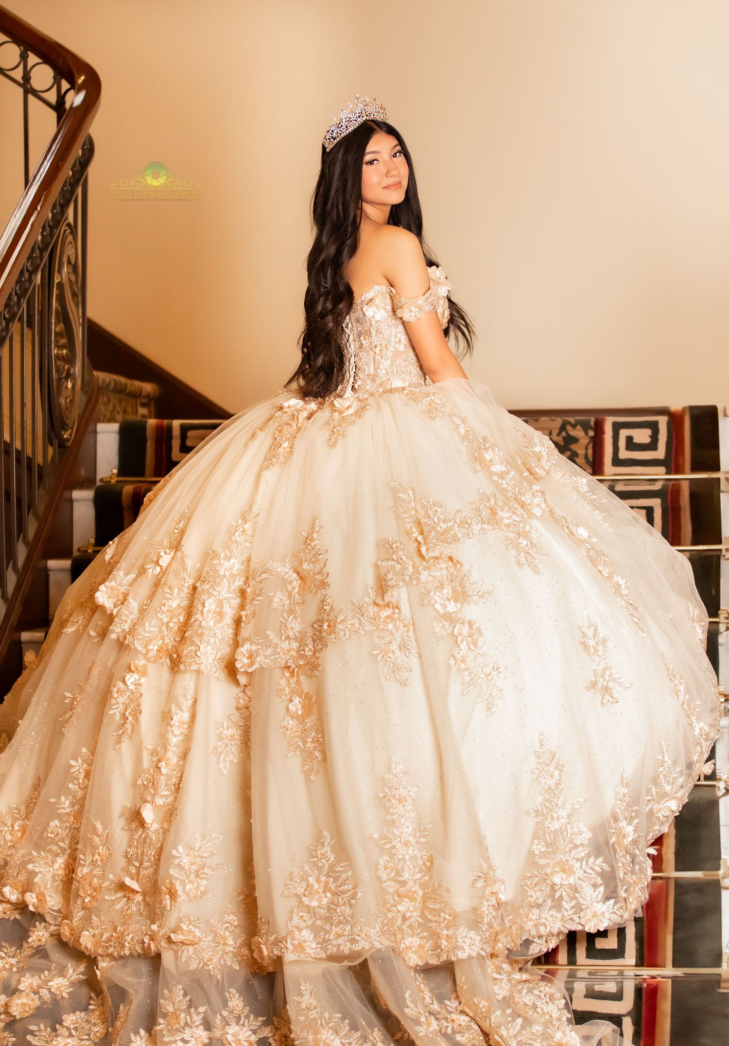 Young woman in a gold floral ball gown with off-shoulder neckline and tiara poses on elegant staircase.