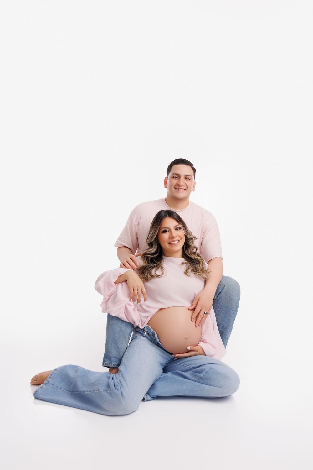 Pregnant woman sitting on floor with partner behind her, both smiling, wearing pink and jeans on white background.