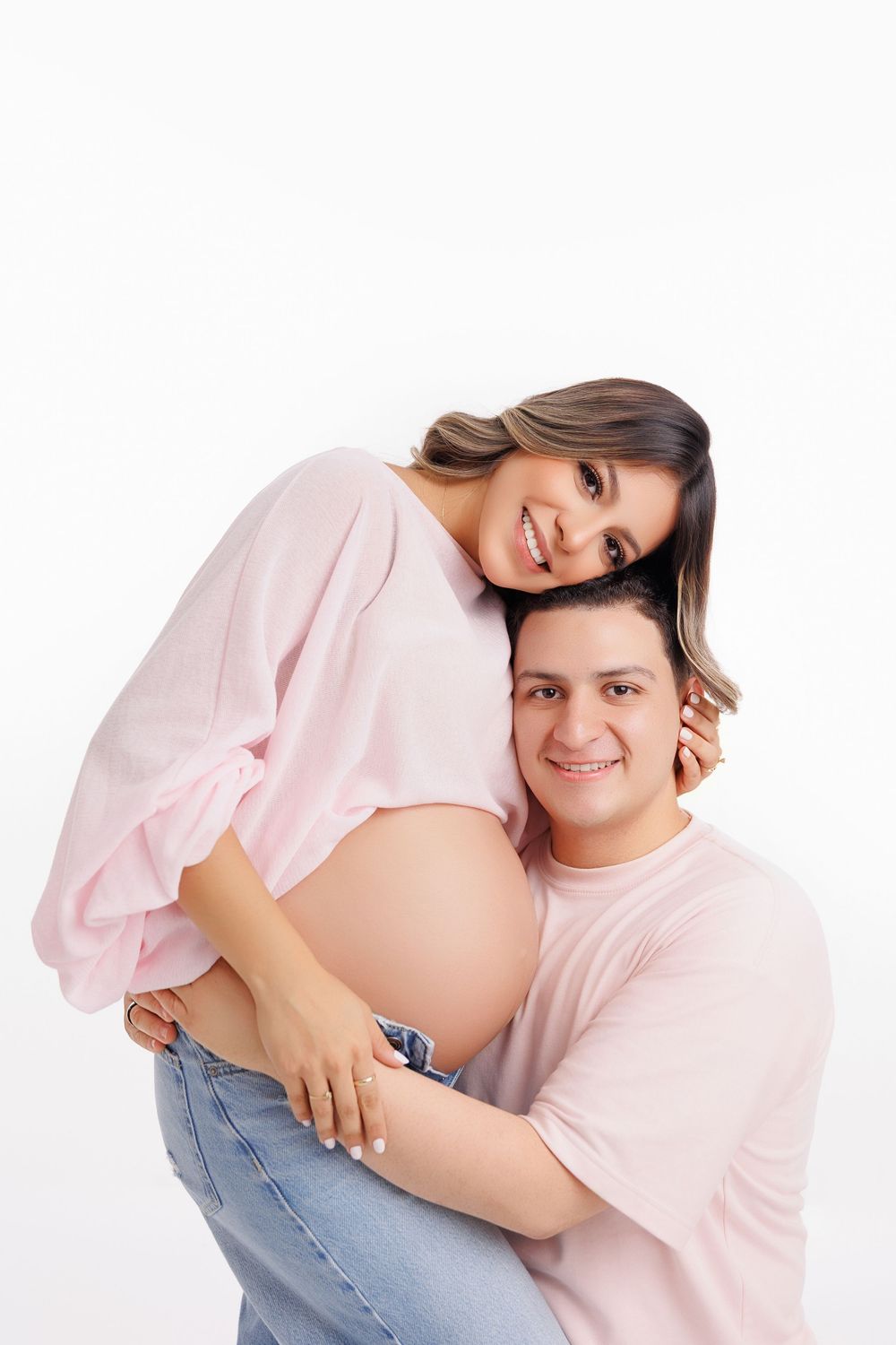 Happy pregnant couple embracing, woman in pink top with baby bump, man hugging her from behind, white background.