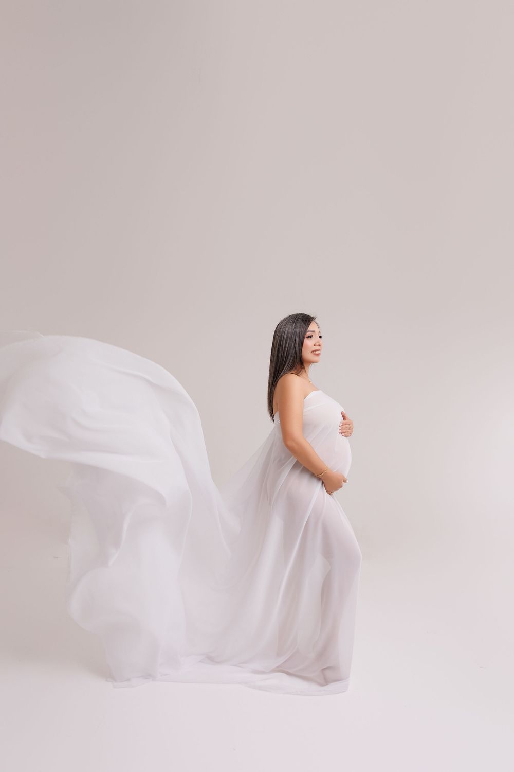 Pregnant woman in flowing white fabric draped elegantly, cradling her baby bump in a serene studio setting.