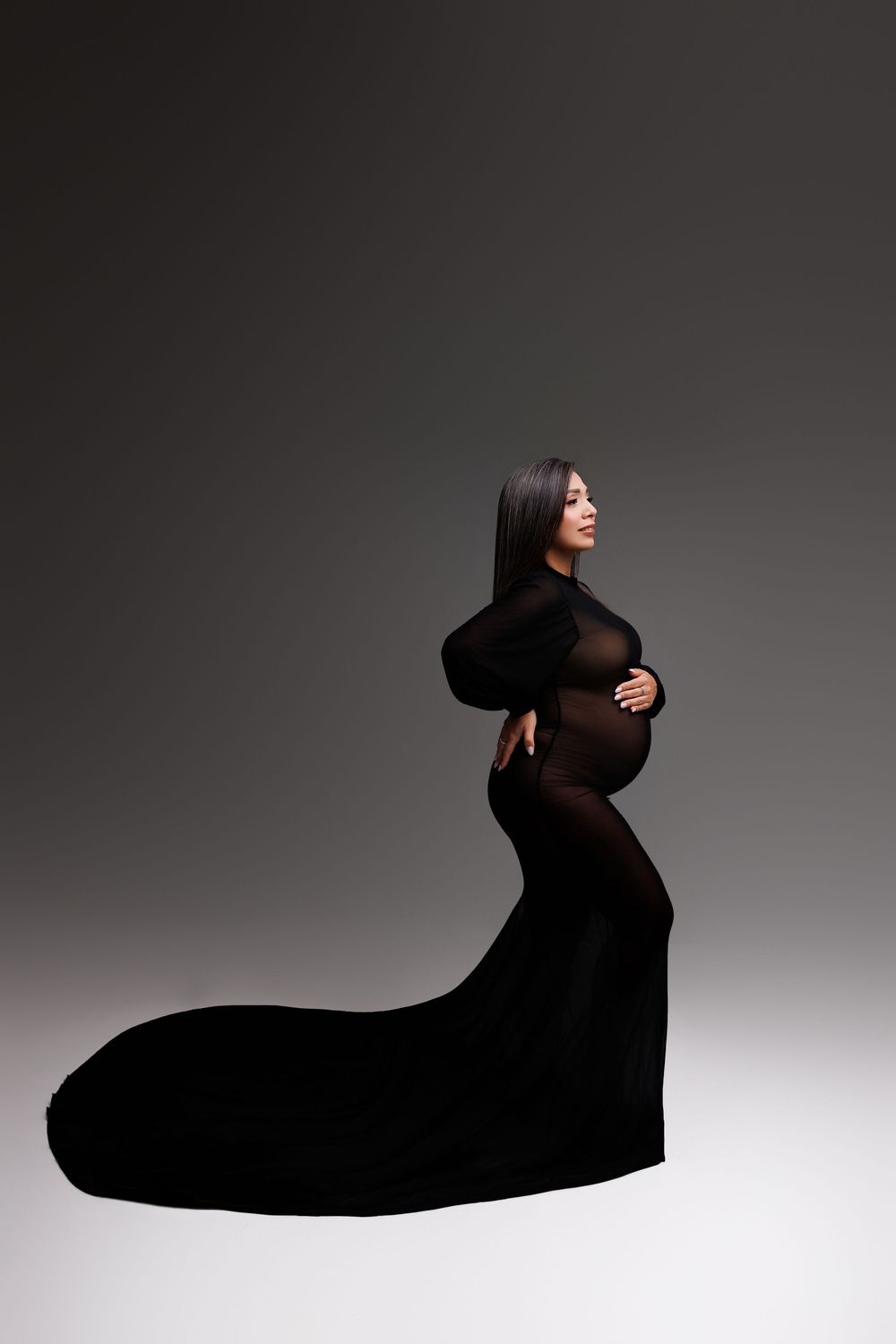 Pregnant woman in elegant black sheer gown with train, posing sideways in studio, cradling baby bump.