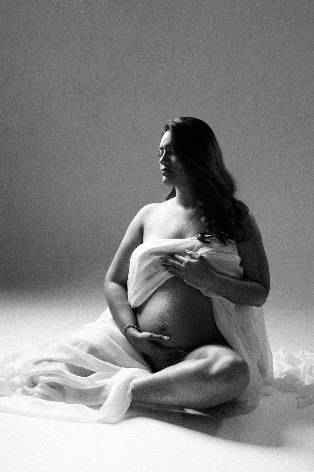 Black and white maternity photo of a pregnant woman sitting gracefully, draped in sheer white fabric, cradling her baby bump.