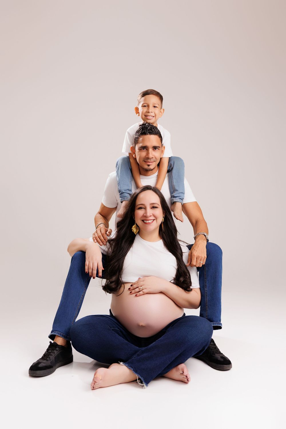 Pregnant woman sits cross-legged with husband and son stacked behind her in matching white tops and jeans for a family photo.